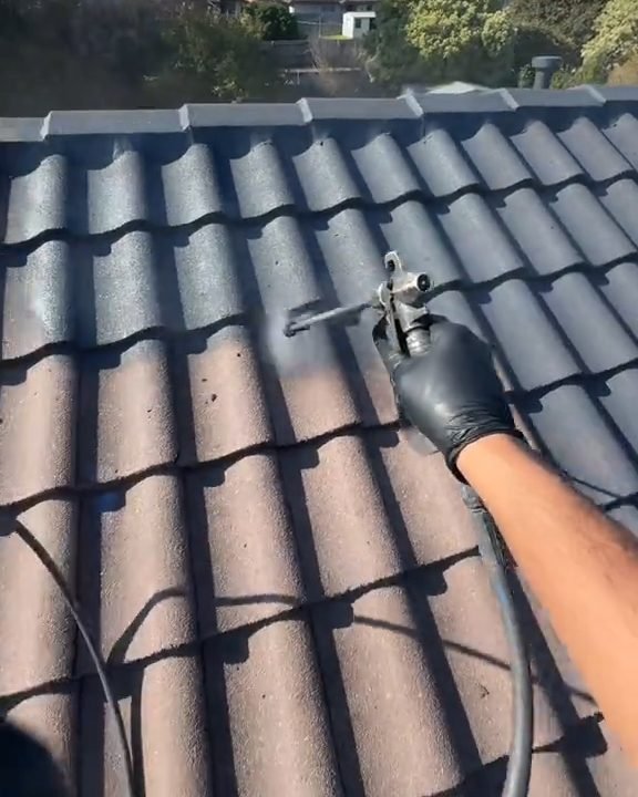 A gloved hand spraying a dark coat onto roof tiles